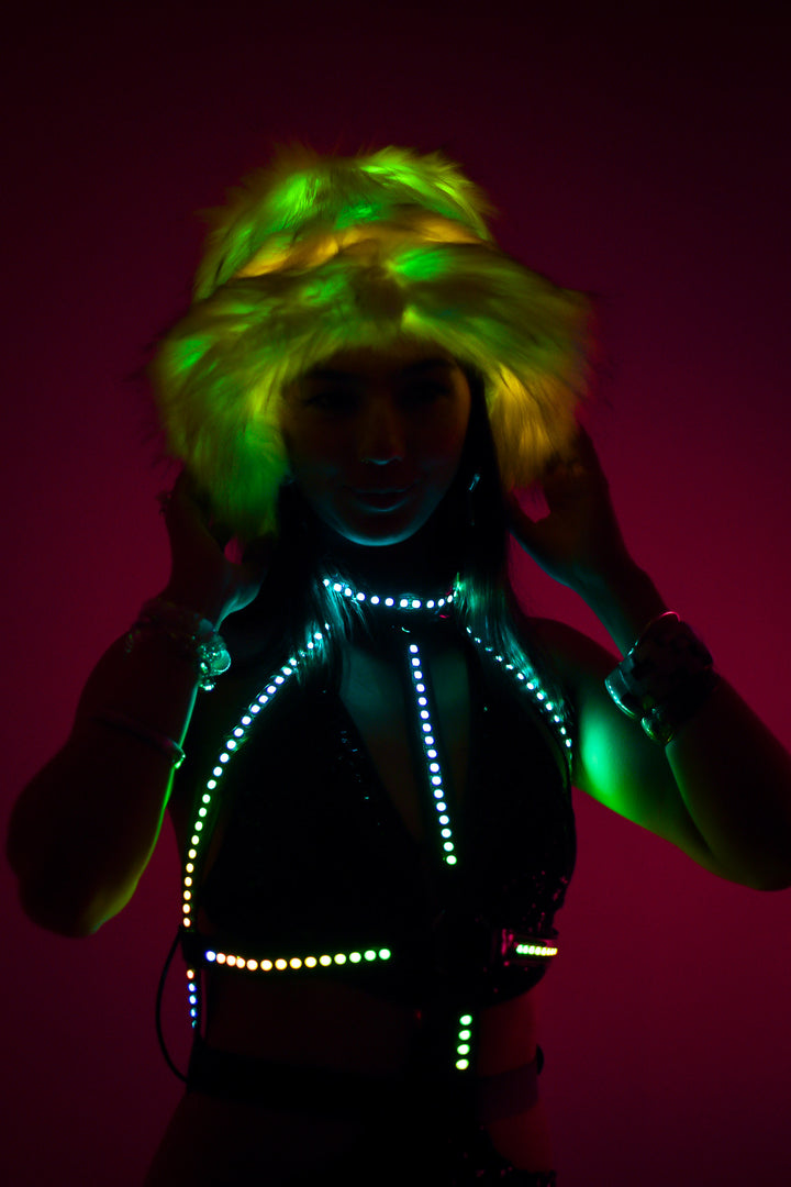 Afterglow Cross LED Harness glowing green and yellow under faux fur hat. Glow Up Bikini Harness for rave and festival looks at Wild Thing.
