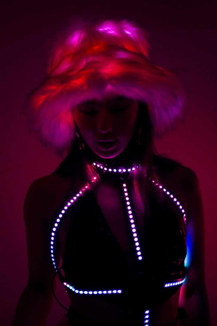 Festival-ready Afterglow Cross LED Harness in pink and purple glow paired with faux fur hat. Glow Up Bikini Harness by Wild Thing.