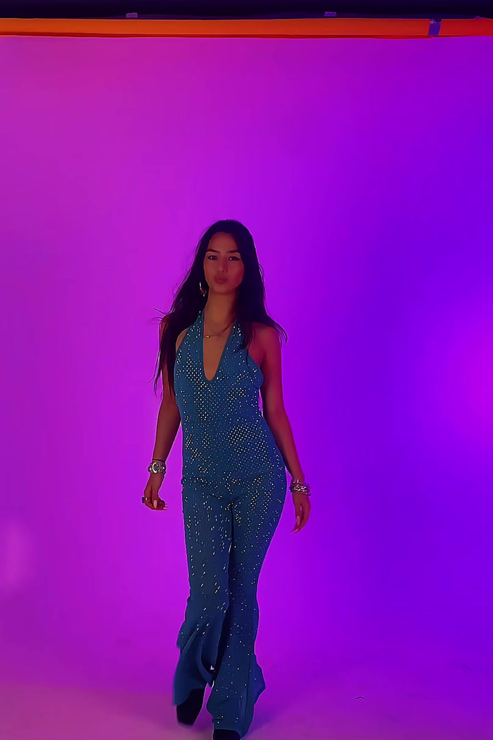 Model strutting in The Disco Tide Catsuit under purple lighting, highlighting its glittery teal fabric and flared silhouette.