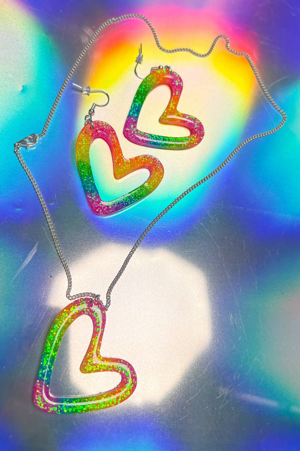 Glittery rainbow heart earrings and necklace on holographic surface